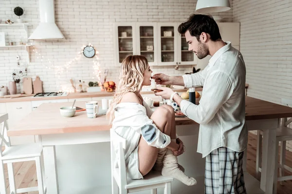 Μελαχρινή γενειοφόρος άνδρας με ένα άσπρο πουκάμισο διατροφή τη σύζυγό του στην κουζίνα — Φωτογραφία Αρχείου