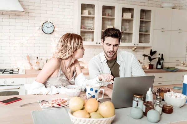 Barbudo bonito homem positivo em uma camisa branca mostrando fotos em um laptop para sua esposa — Fotografia de Stock