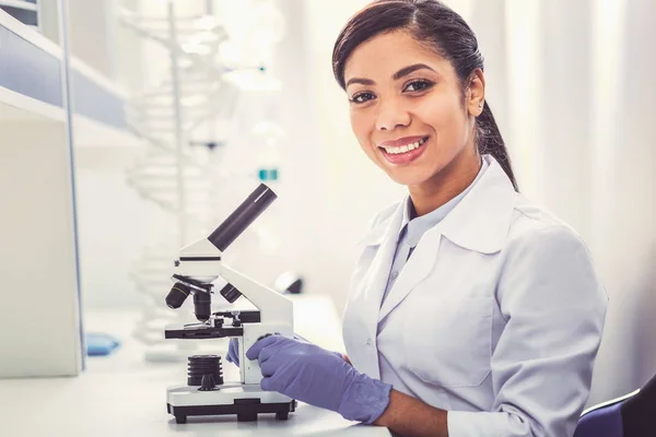 Young chemist wearing white uniform sitting near microscope