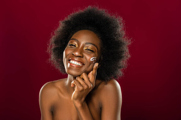 For skin. Delighted afro American woman smiling while putting cream on her cheek