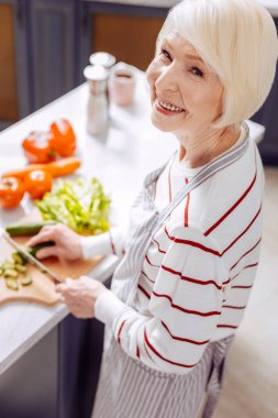 Pişirme büyükanne. Gülümseyen ve modern mutfak ve kesme sebze dururken memnun seyir neşeli duygusal kadın kıdemli