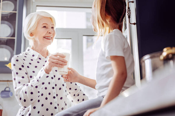 Cheerful aged woman smiling while giving glass of milk to child