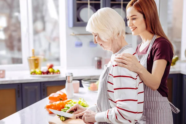 Mosolyogva idősebb nő főzés és megható vállán lánya — Stock Fotó