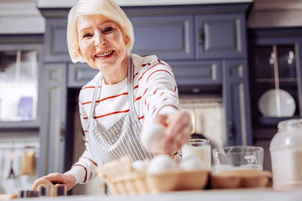 焼きます 陽気の親切な年配の女性は笑みを浮かべて キッチンでおいしい料理を調理しながらボックスから卵を取って — ストック写真