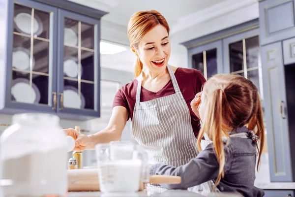 Excited woman looking at her daughter and smiling while cooking with her