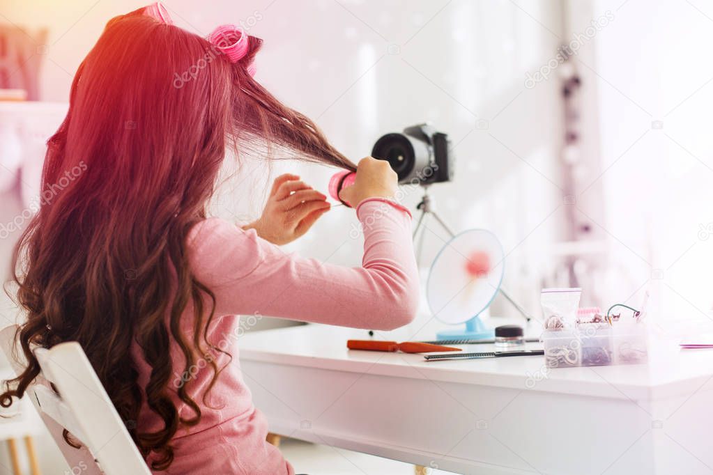 Modern little girl filming her life blog while using pink hair curlers making hairstyle