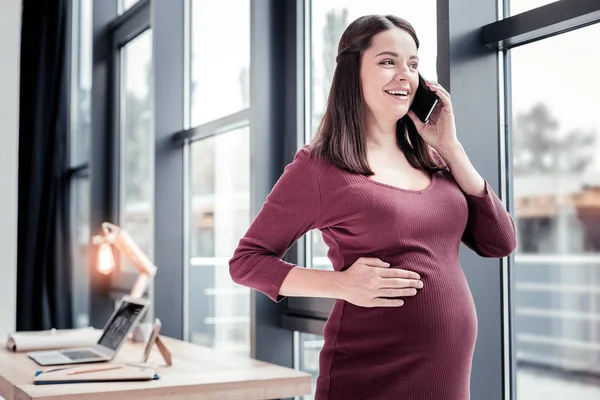 Joven encantadora mujer riendo mientras habla en el teléfono de pie cerca de la ventana — Foto de Stock