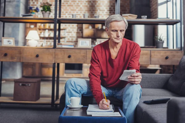 Starszy mężczyzna robiąc notatki, siedząc w swoim pokoju gościnnym — Zdjęcie stockowe