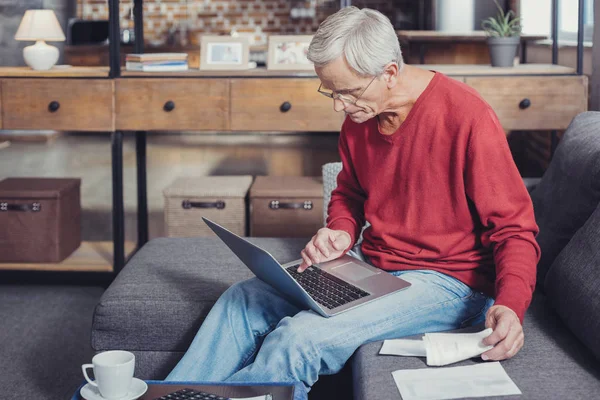 Внимательный пенсионер держит в руках ноутбук и оплачивает счета онлайн — стоковое фото