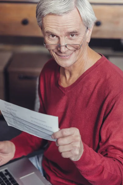 Pensionista positivo sonriendo mientras sostiene una factura y una computadora portátil — Foto de Stock