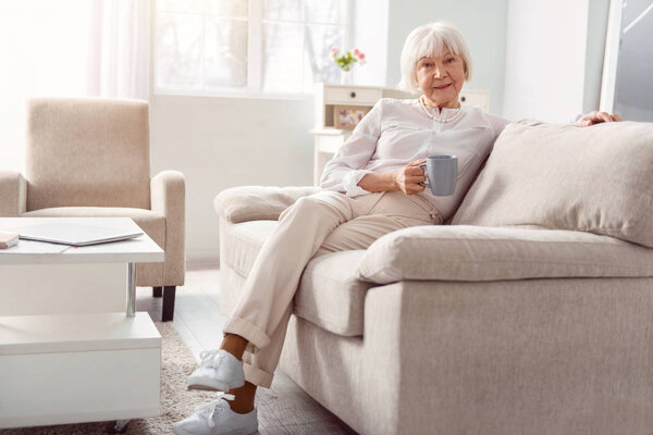 Charming elderly lady relaxing with coffee cup on couch