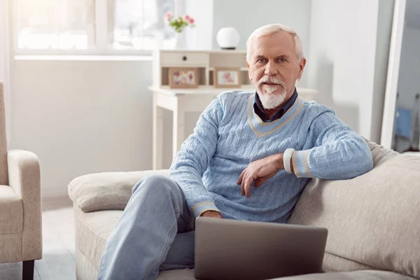 Обаятельный пожилой человек позирует, используя ноутбук на диване — стоковое фото