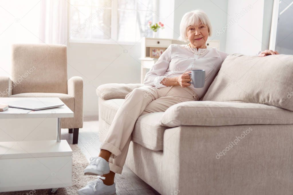 Charming elderly lady relaxing with coffee cup on couch