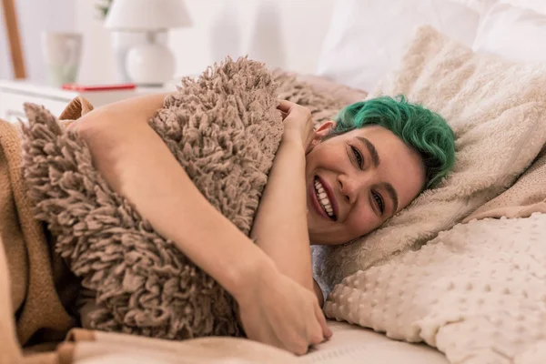 Cheerful woman hugging little fluffy pillow while chilling in bed