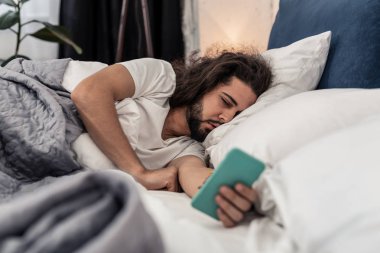 Sleepy brunette man looking at his smartphone screen clipart