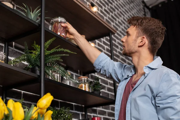 Schöner junger bärtiger Mann stellt Glas mit Gewürzen ins Regal — Stockfoto