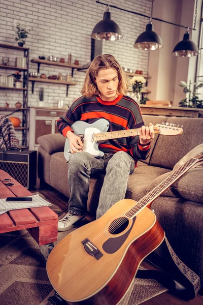 Estudiante vistiendo jeans tocando la guitarra sentado en el sofá — Foto de Stock