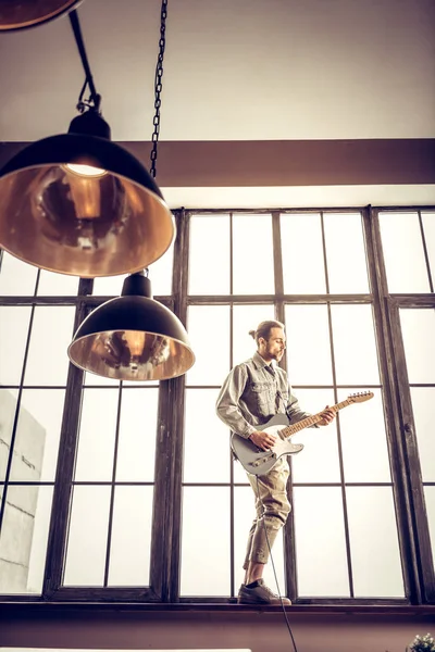 Rock musiker bär jeansskjorta spela gitarr nära fönster — Stockfoto