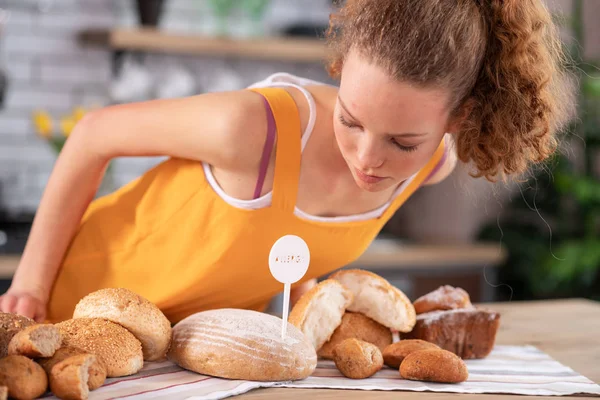 Femeia curioasă cu părul creț în șorț galben observând o grămadă de pâine — Fotografie, imagine de stoc