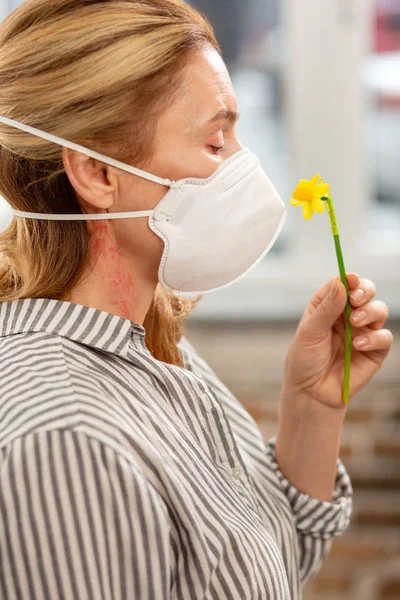 Mujer rubia usando máscara mientras huele pequeña flor amarilla — Foto de Stock