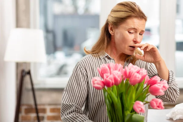 Vrouw permanent in de buurt van het venster met bloemen niezen met allergie — Stockfoto