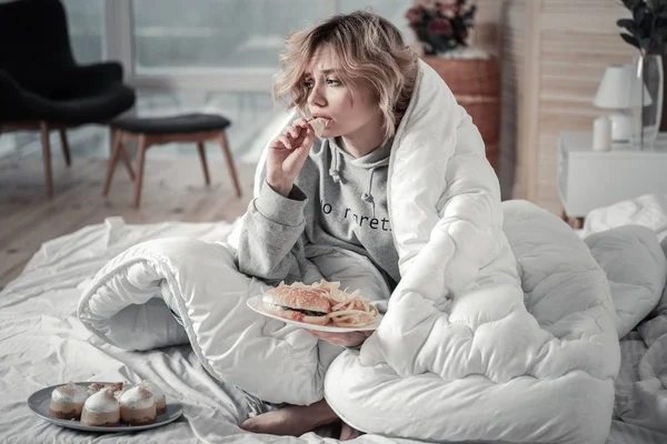Üzgün ve yalnız kadın yatakta hamburger ve patates kızartması yemek — Stok fotoğraf
