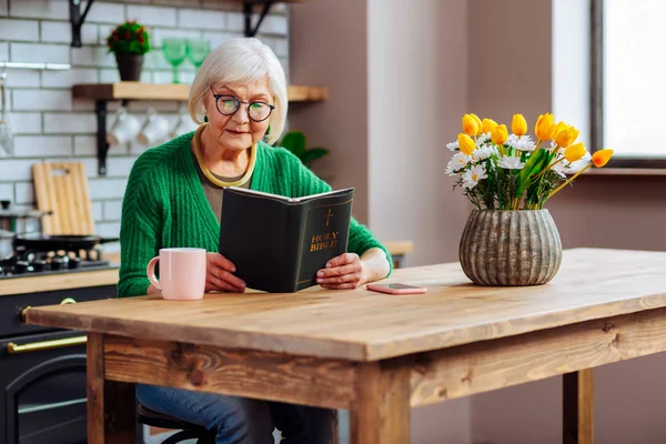 Dama encantadora com cabelo prateado leitura Bíblia na mesa da cozinha — Fotografia de Stock