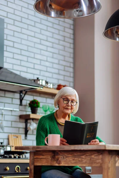 Idős nő elfoglalva, szem előtt tartva olvasata a Biblia konyha — Stock Fotó