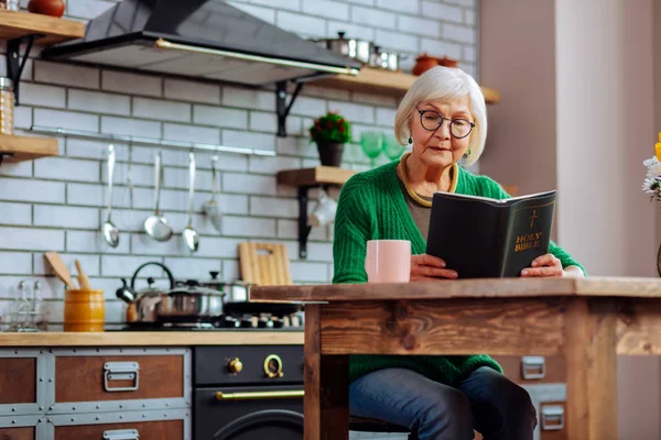 Régi asszony élvez olvasó a bibliai könyv a konyhaasztalon — Stock Fotó