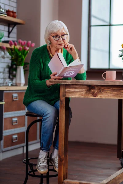 Стареющая женщина поглощена книгой, сидя на меблированной кухне — стоковое фото