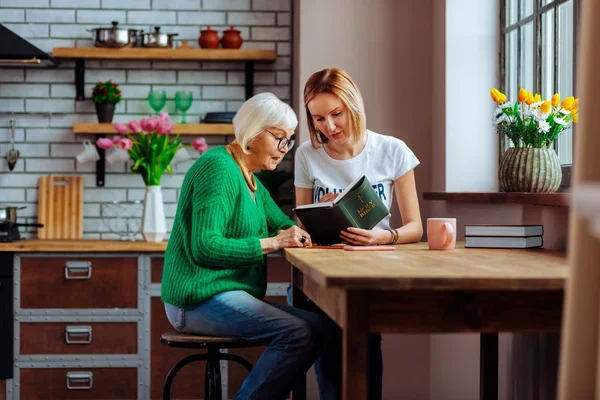 Joven voluntario de la iglesia leyendo la Biblia con pensionista en casa cocina — Foto de Stock