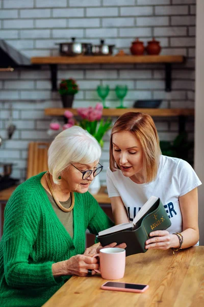 Bewitching dame sitting at table and reading Bible with lady — Stock Photo, Image