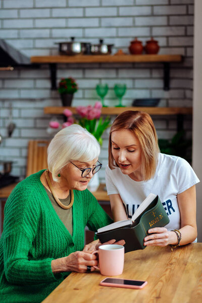 Bewitching dame sitting at table and reading Bible with lady