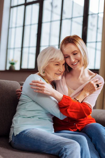 Charming woman affectionately cuddling elderly madam sitting on couch — Stock Photo, Image