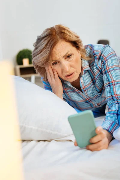 Mulher de cabelos claros expressiva vestindo pijama quadriculado e inspecionando smartphone — Fotografia de Stock