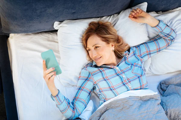 Smiling positive adult lady lying on her back and holdying mobile phone — Stock Photo, Image