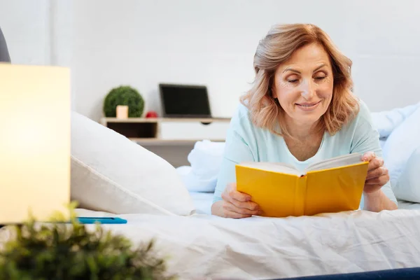 Положительная улыбающаяся женщина лежит на кровати и читает книгу в твёрдом переплете — стоковое фото