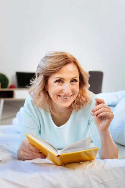 Neşeli ışık saçlı kadın yatağında dinleniyor ve ilginç bir kitap okuma — Stok fotoğraf