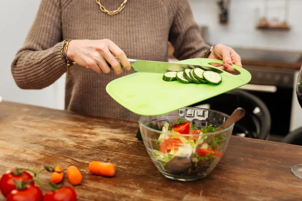 Hard-working accurate woman adding chopped cucumbers into glass bowl — Stock Photo, Image