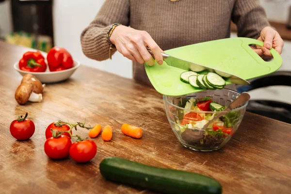 Attentive woman in beige sweater throwing vegetables into glass bowl — Stock Photo, Image