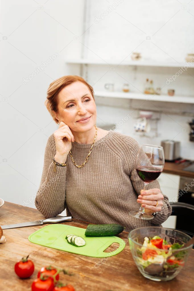 Beaming good-looking lady holding glass of wine while leaning on table