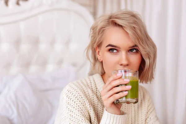 Blonde-haired woman drinking healthy green juice in bedroom — Stock Photo, Image