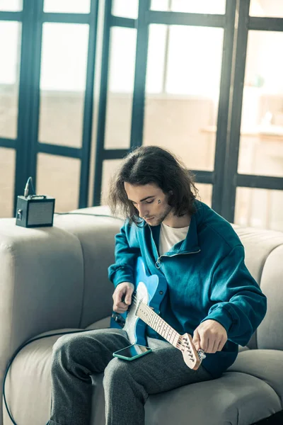 Chico guapo de pelo oscuro tonificando su guitarra electrónica y comprobando las redes sociales — Foto de Stock