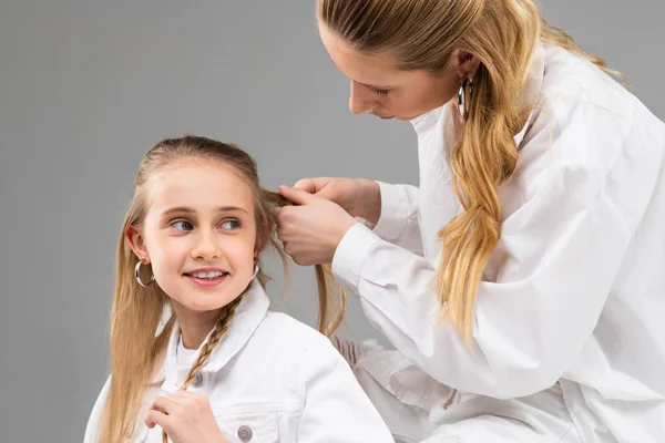 Внимательная взрослая женщина в белой рубашке собирает волосы своей младшей сестры — стоковое фото