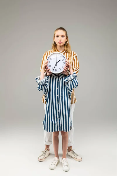 Little girl in oversize striped shirt closing her face with round clock — Stock Photo, Image