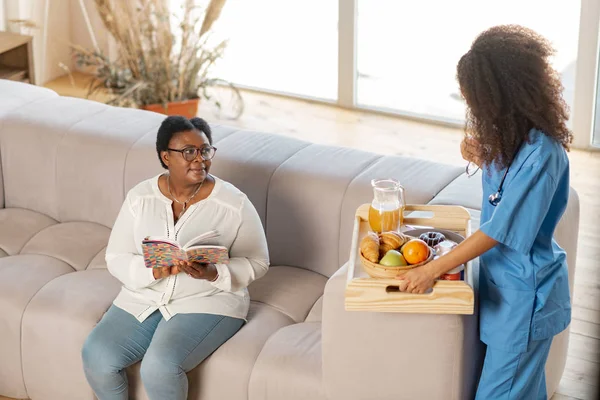 Enfermera trayendo croissants y frutas para anciana sentada en sofá — Foto de Stock