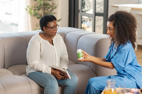 Curly dark-skinned nurse giving pack with pills to sick woman — Stock Photo, Image