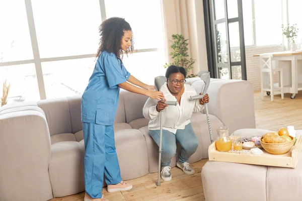 Медсестра помогает женщине встать с дивана с костылями — стоковое фото