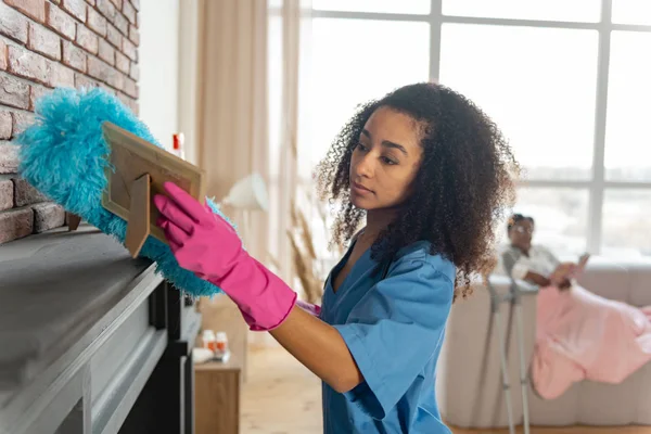 Private nurse looking at photo frame while dusting the fireplace — Stock Photo, Image