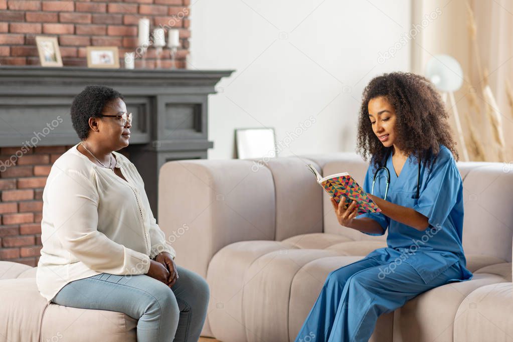Medical attendant sitting in the living room and reading book for patient